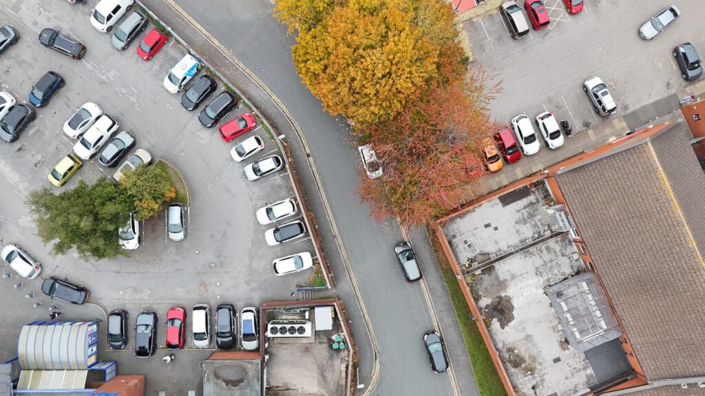 Drone view of a town-centre car park with angled bays following a curved boundary, trees and a neighbouring road
