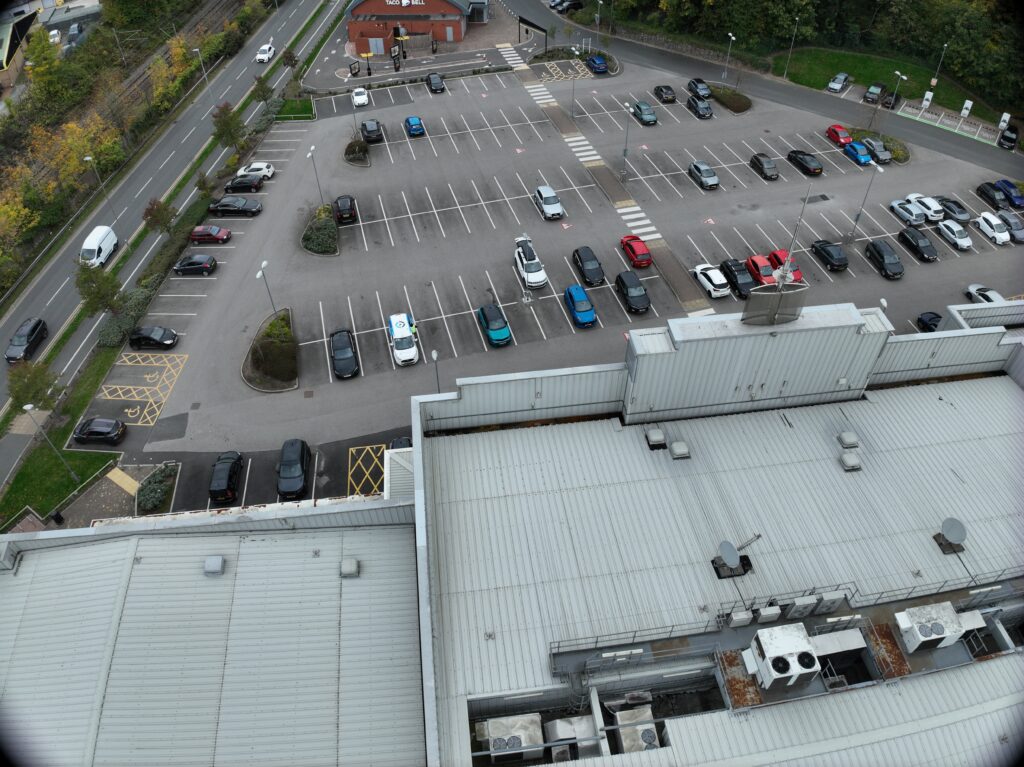 Oblique drone view of a large retail car park with EV charging bays, pedestrian crossings and the building roof in the foreground
